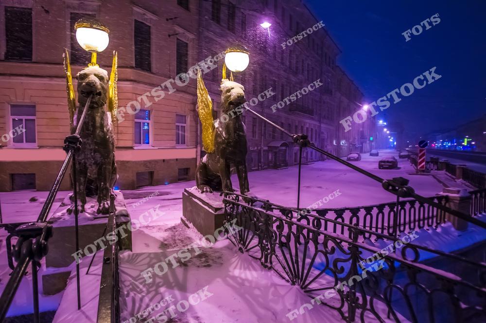 На фотографии изображены скульптуры грифонов на Банковском мосту в Санкт-Петербурге, покрытые снегом. Вечернее освещение подчеркивает золотые крылья статуй, создавая атмосферу загадочности и уюта. Снегопад и ночная подсветка добавляют композиции зимнюю романтику, подчеркивая красоту исторического центра Петербурга. Это место привлекает внимание туристов в любое время года, особенно в зимние месяцы.